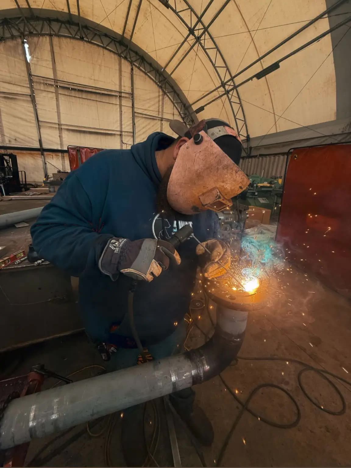 A SWAT welder grinding a pipe joint, sparks flying, inside a fabric-roofed shop